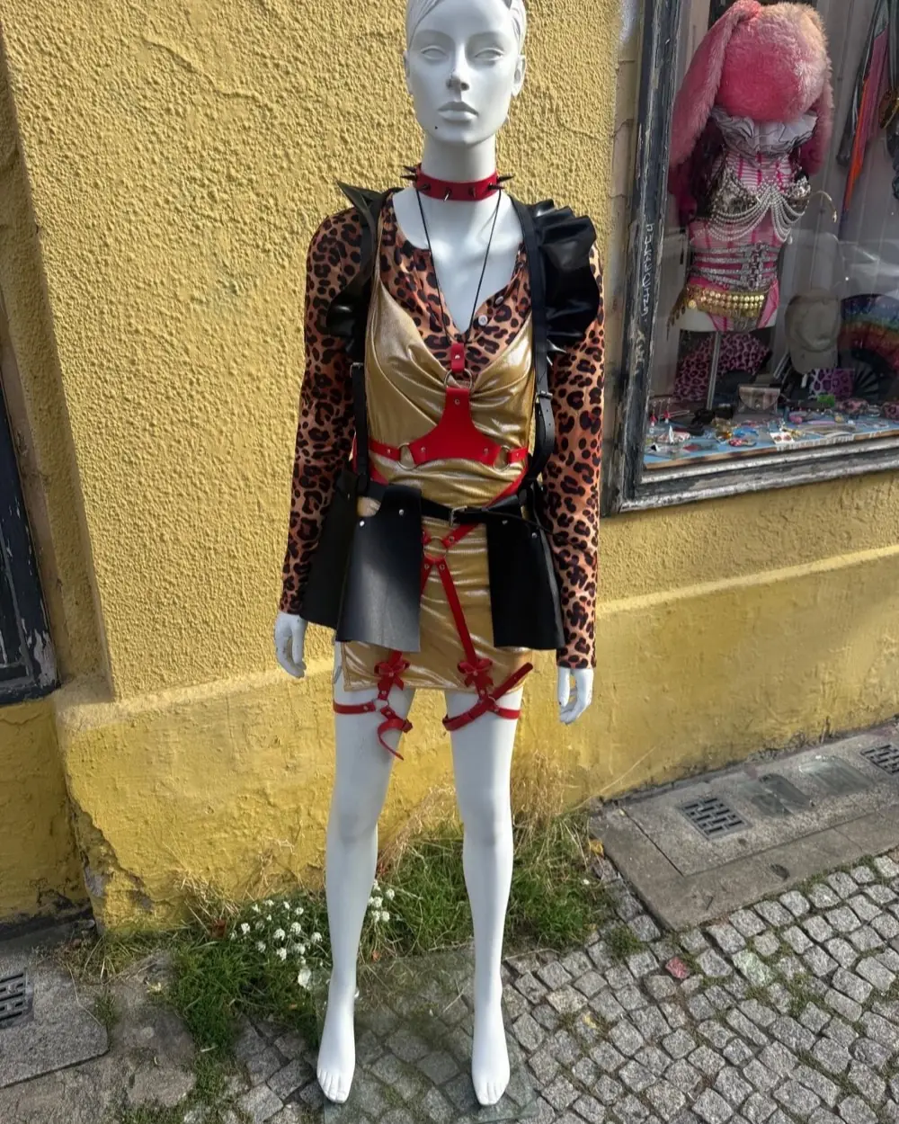 Puppe vor gelber Hauswand in Leo-Shirt, Goldkleid und roten Lederharness